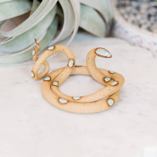 Speckled tan ceramic snake with mint green spots coiled on marble, head raised, air plant behind
