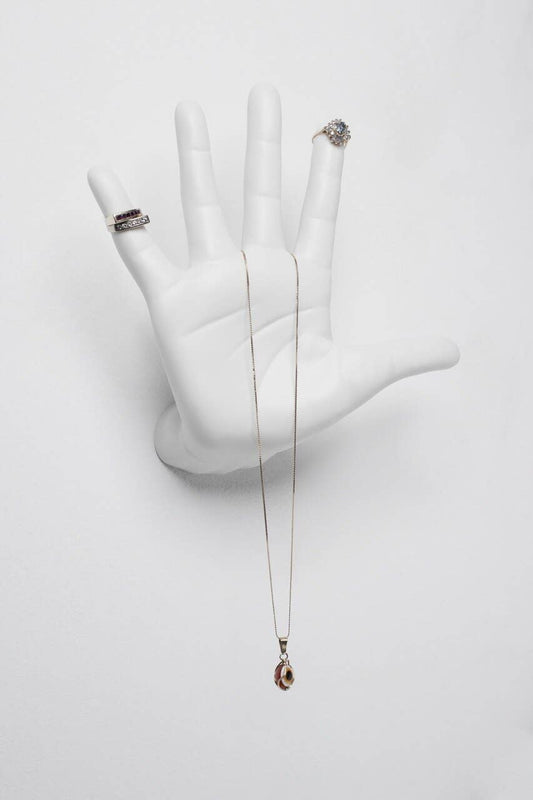 White ceramic open-palm hand mounted on a white wall, used as a jewelry holder for rings and a pendant