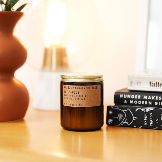 No. 32 Sandalwood Rose Soy candle in amber jar with craft label and brass lid. On table next to books and vase.