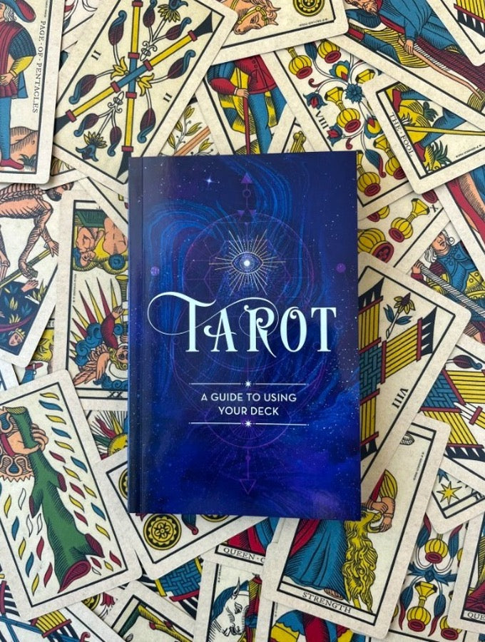 Tarot guidebook with a navy cosmic cover, A guide to using your deck, on a pile of Marseilles tarot cards