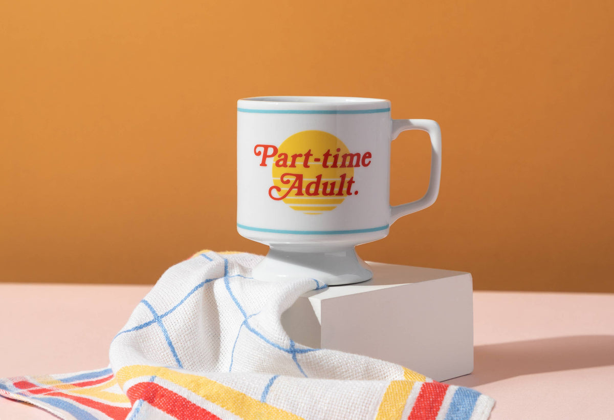 Part-time Adult pedestal mug on a white block with a striped tea towel against an orange wall