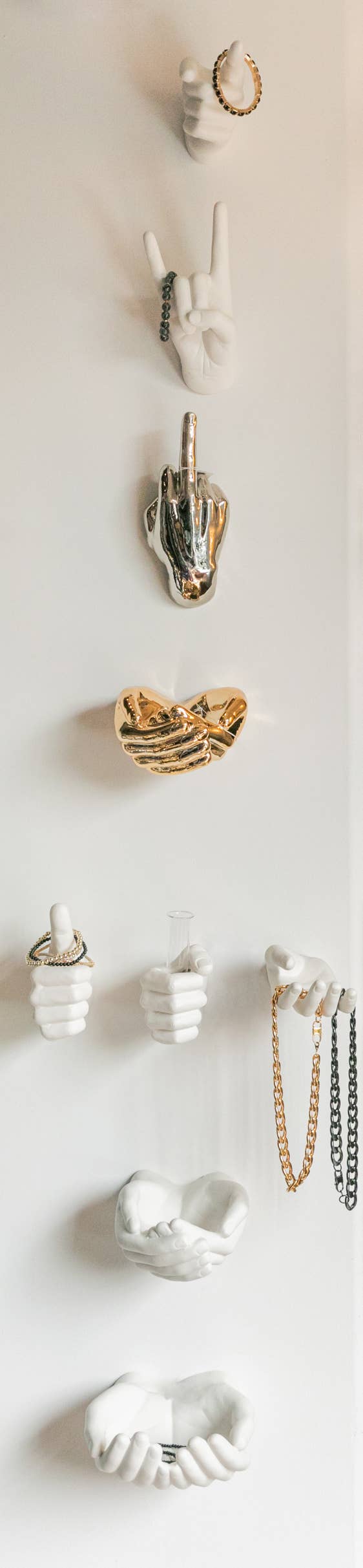 Wall display of white, gold, and silver hand sculptures holding rings, bracelets, necklaces, and a vase tube