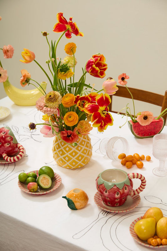 Pineapple vase holding orange and red tulips and ranunculus on a fruit-themed dining table