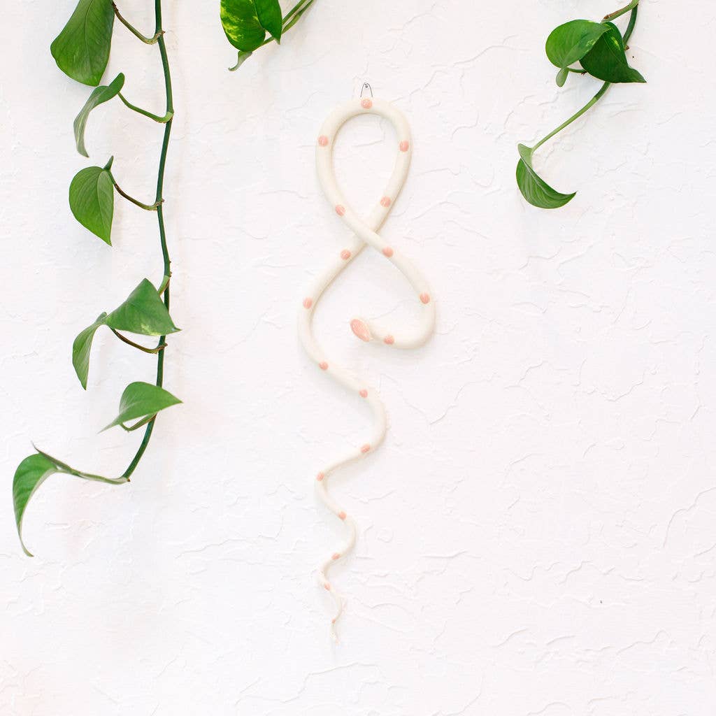 Cream ceramic wall snake with soft pink dots hanging on a white wall framed by pothos leaves