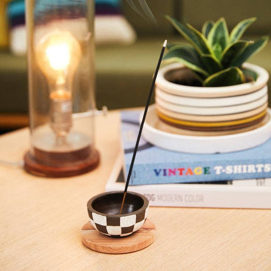 Golden Coast incense stick burning in a black and white checkered holder on a wood table by a plant