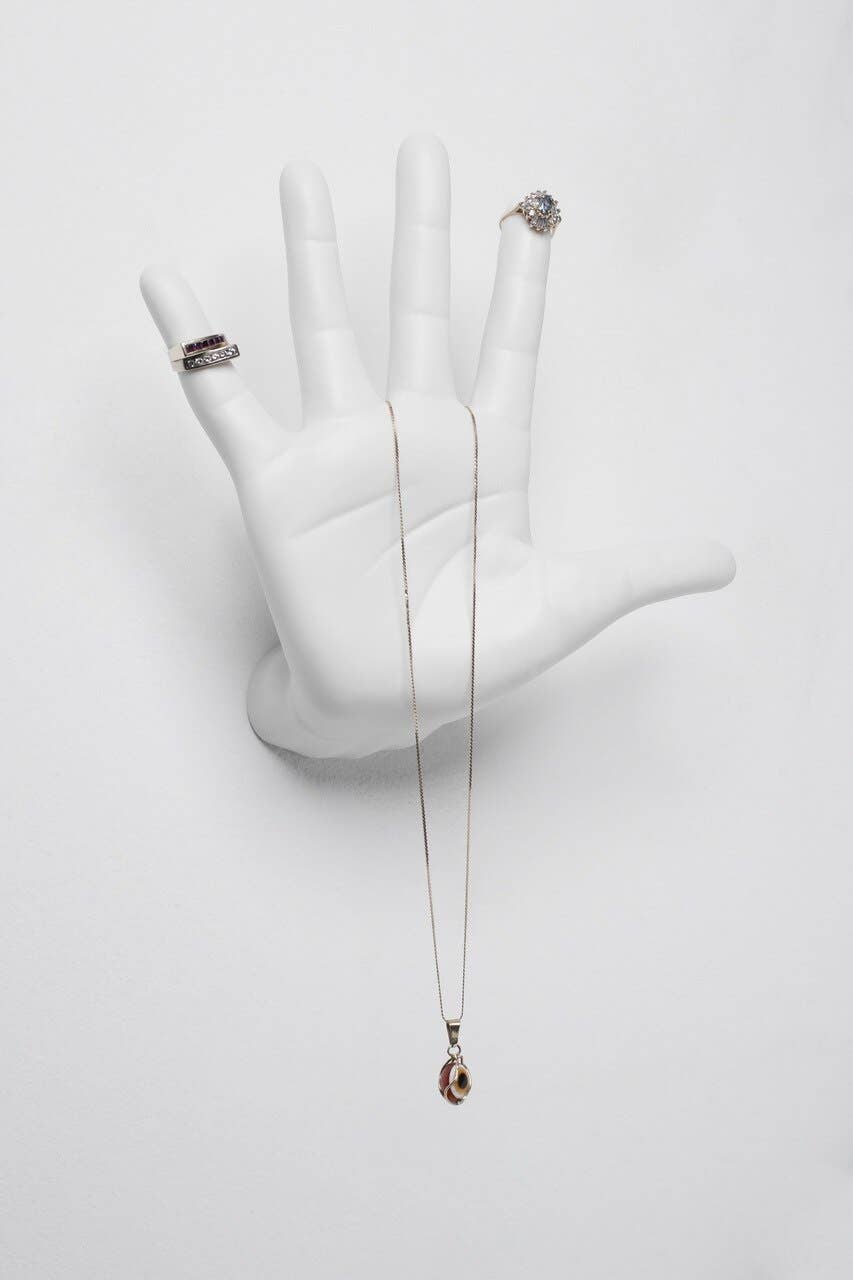 White ceramic open-palm hand mounted on a white wall, used as a jewelry holder for rings and a pendant