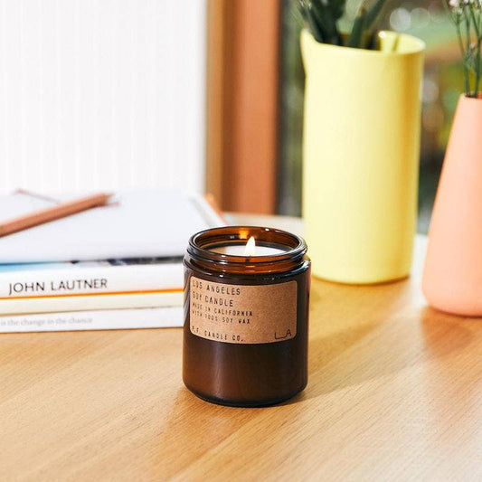Lit Los Angeles soy candle in amber jar on a wood table beside books and yellow and pink vases