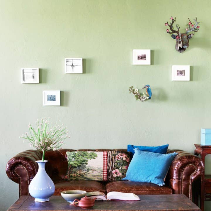 Eco deer head hung on a sage green wall above a leather sofa, with framed photos and a bird