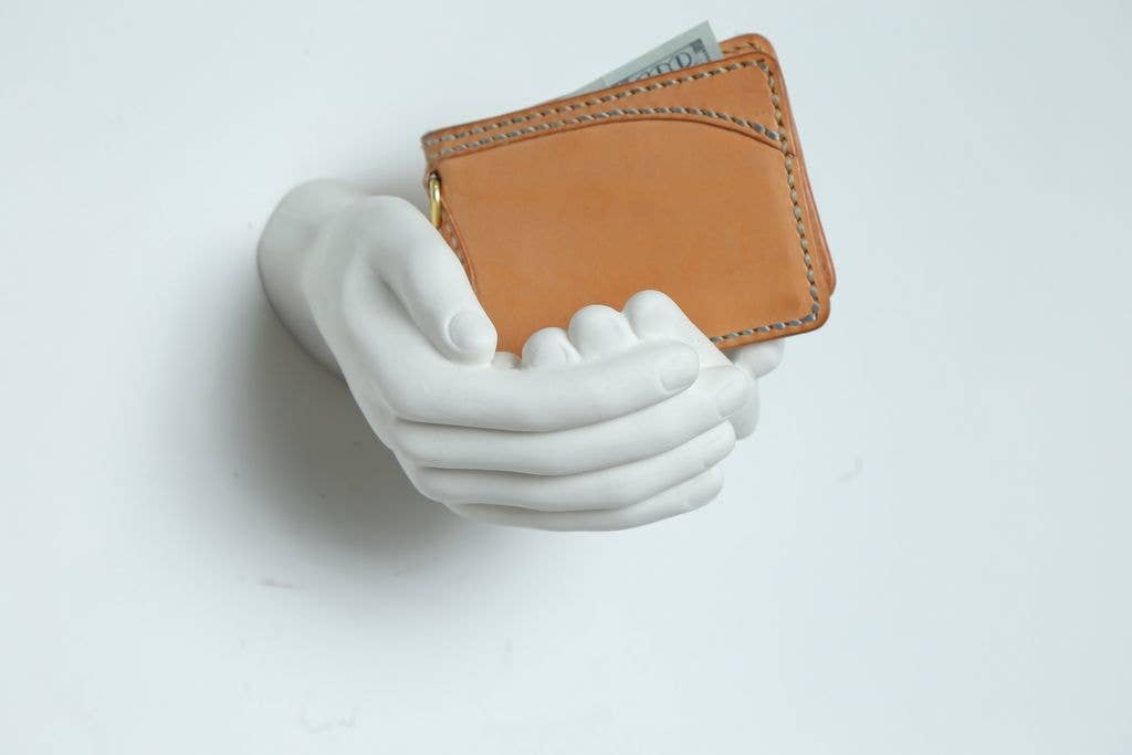 Close-up of white criss-cross hands wall hook holding a tan leather wallet against a white wall