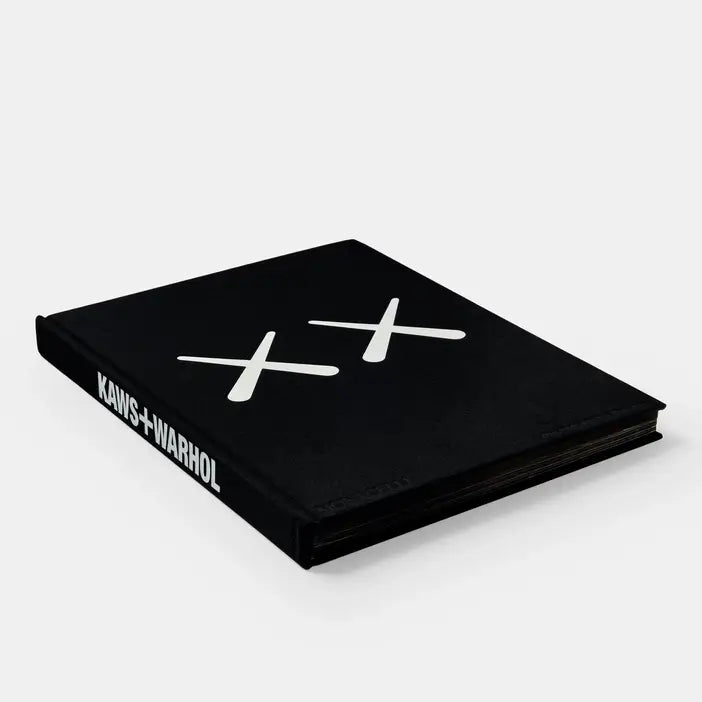 Kaws + Warhol book lying flat at an angle, black cover with white X marks and white spine lettering