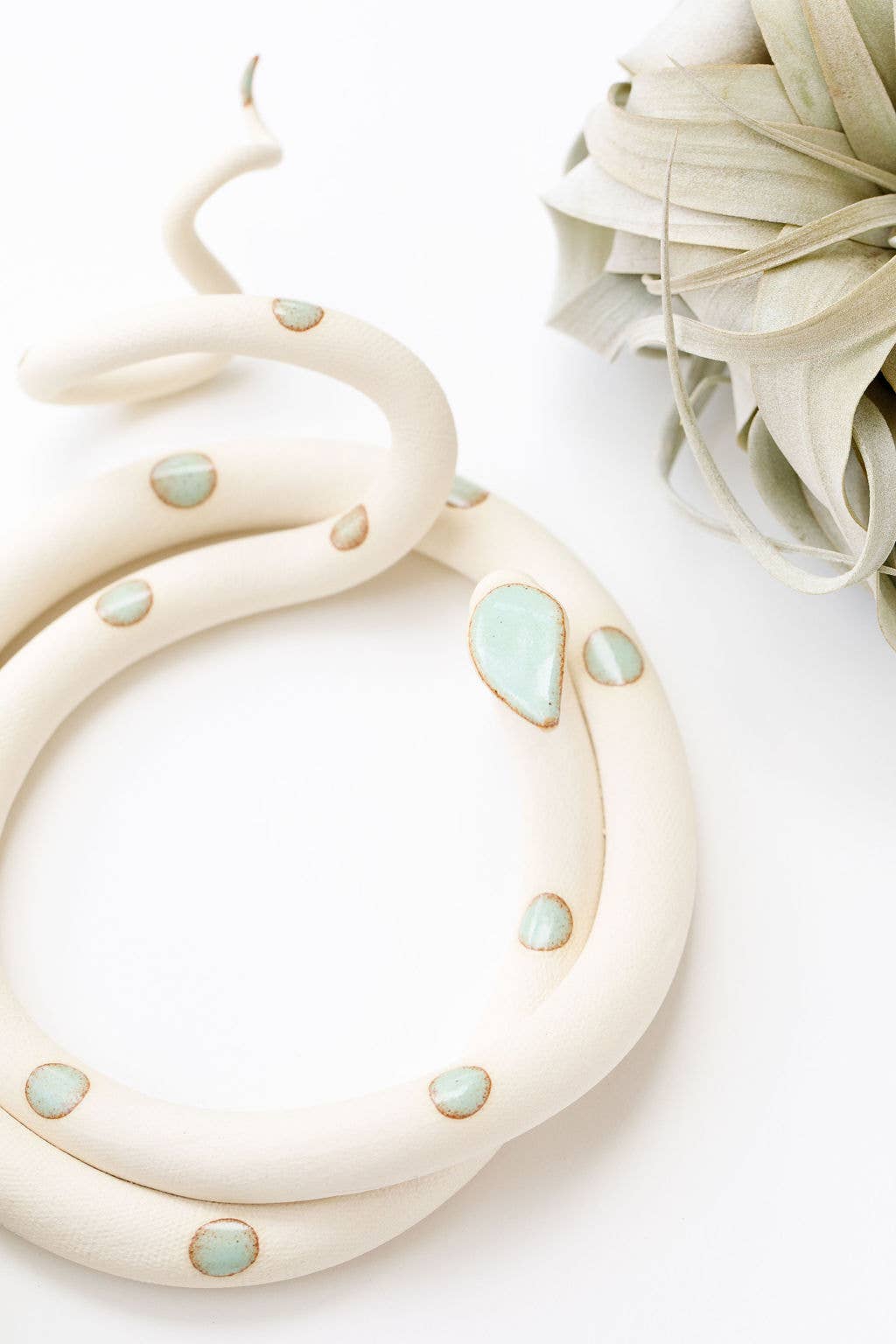 Close-up of a cream ceramic snake with mint green spots and a curled tail, beside an air plant