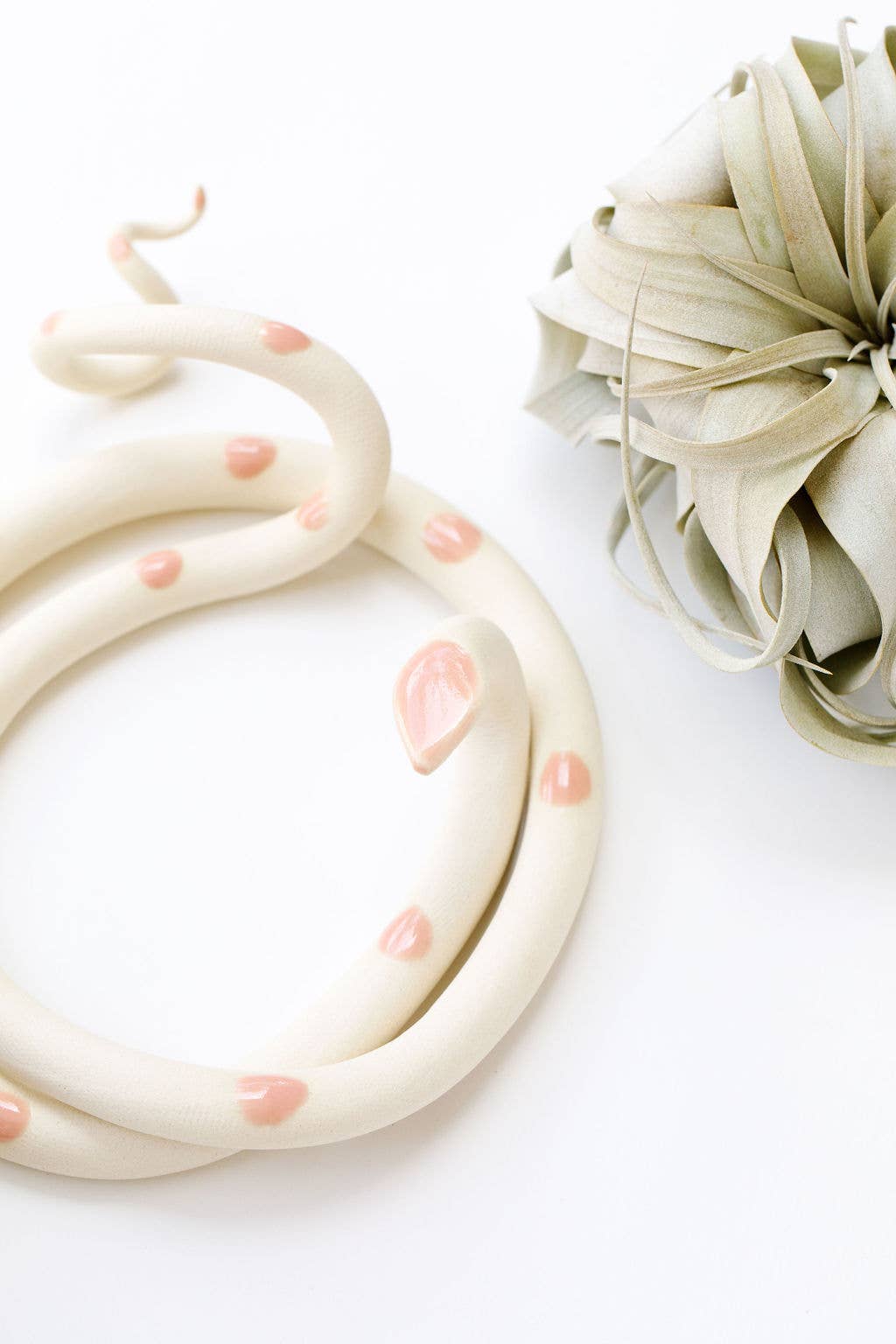 Close-up of a cream ceramic snake with glossy pink spots and a pink head beside an air plant