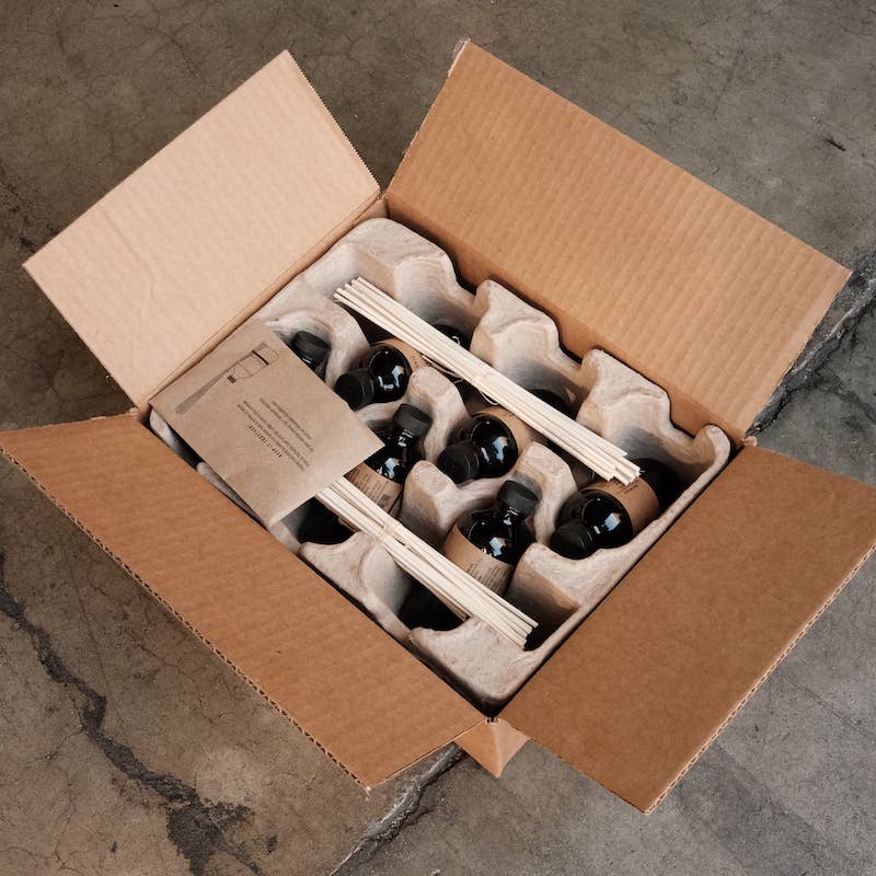 Open shipping box from above holding dark diffuser bottles and bundled reeds in a pulp insert