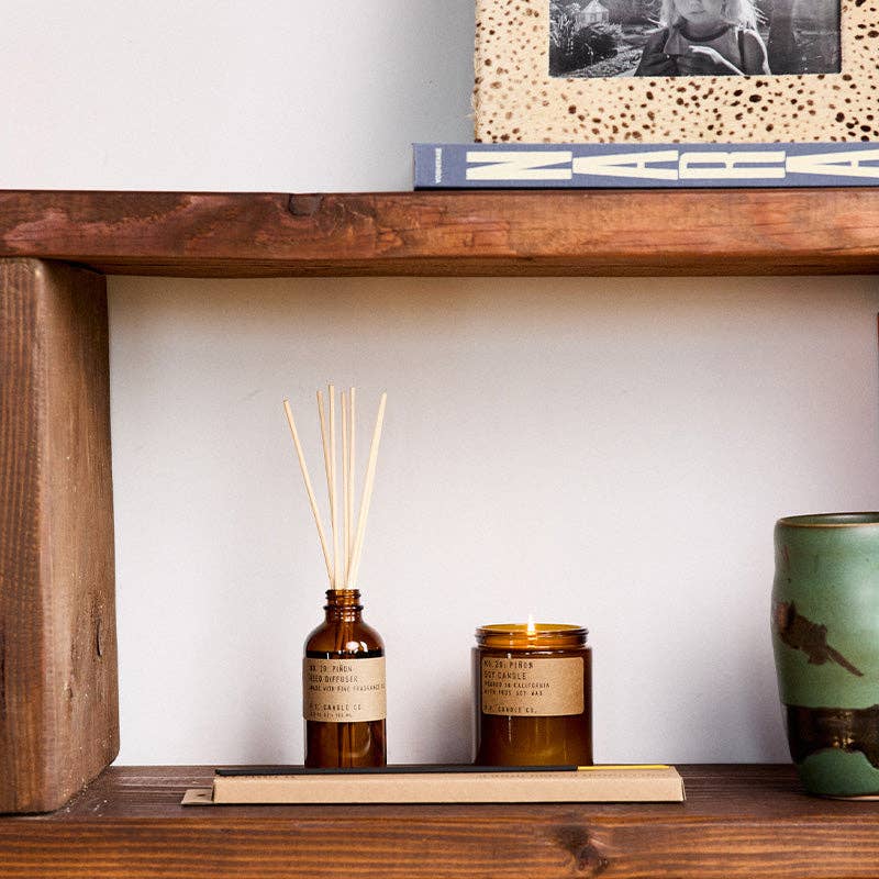 Piñon reed diffuser and lit jar candle on a rustic wood shelf, set on a kraft incense box