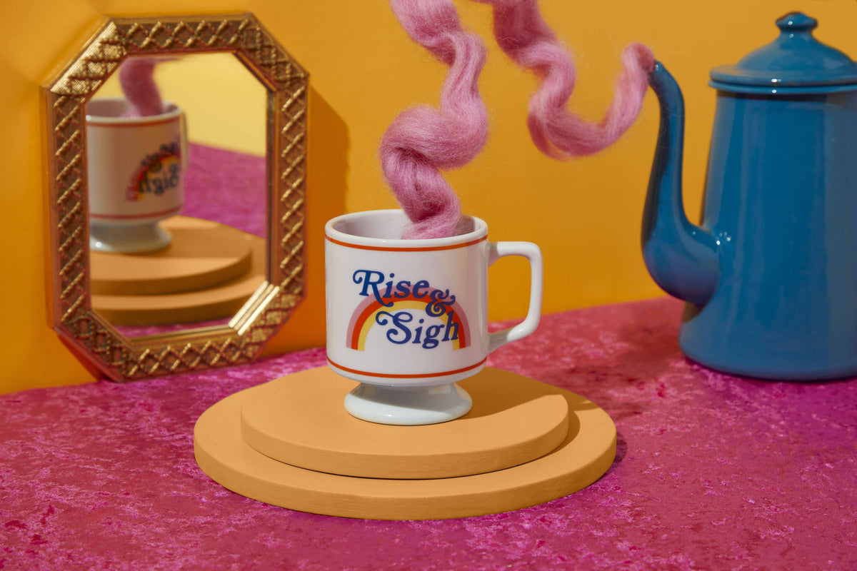 Rise & Sigh pedestal mug on mustard risers with a pink yarn steam swirl, gilt mirror and blue kettle