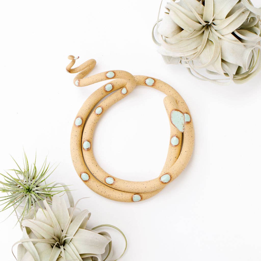 Coiled speckled tan ceramic snake with mint green spots, shown from above beside air plants