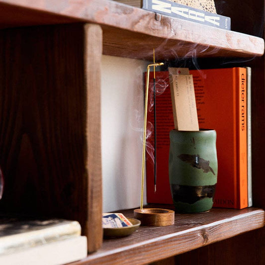 Piñon incense stick burning in a brass holder on a wood shelf beside a green ceramic cup and orange books