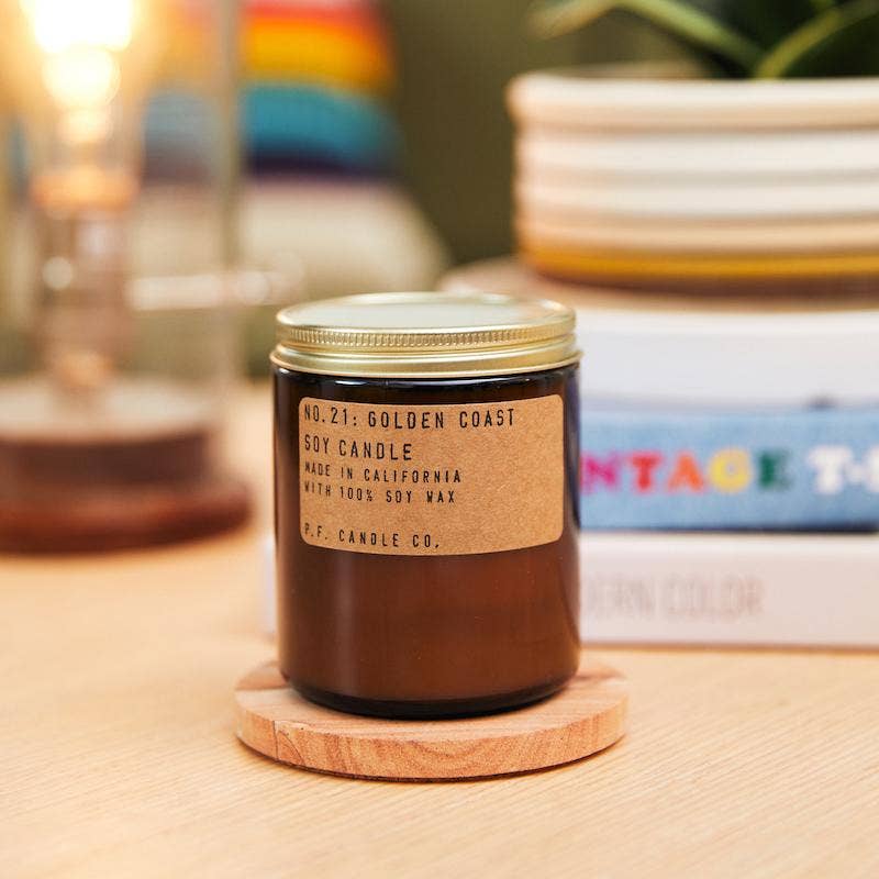Golden Coast soy candle on a wood coaster on a light table, with books and a lamp blurred behind