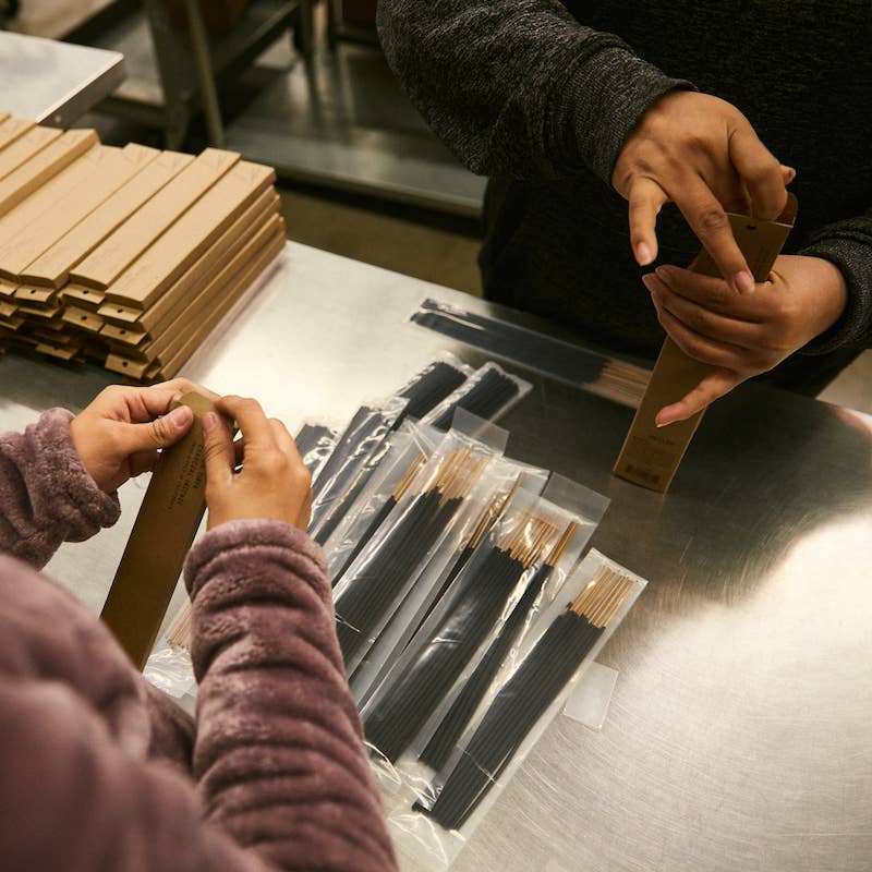 Two people packing black incense sticks into kraft boxes at a stainless steel work table