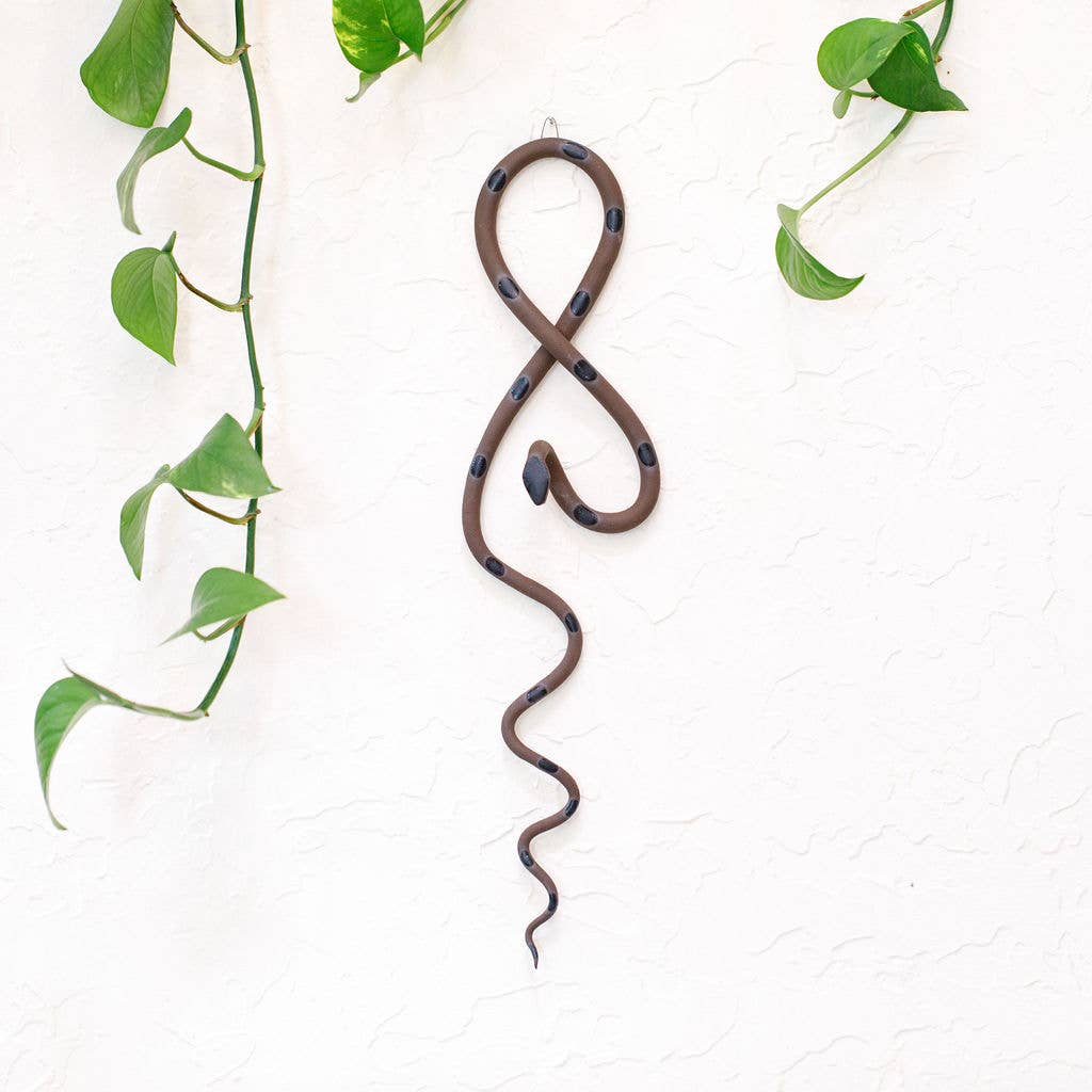 Brown ceramic wall snake with black spots hanging on a white wall between trailing pothos