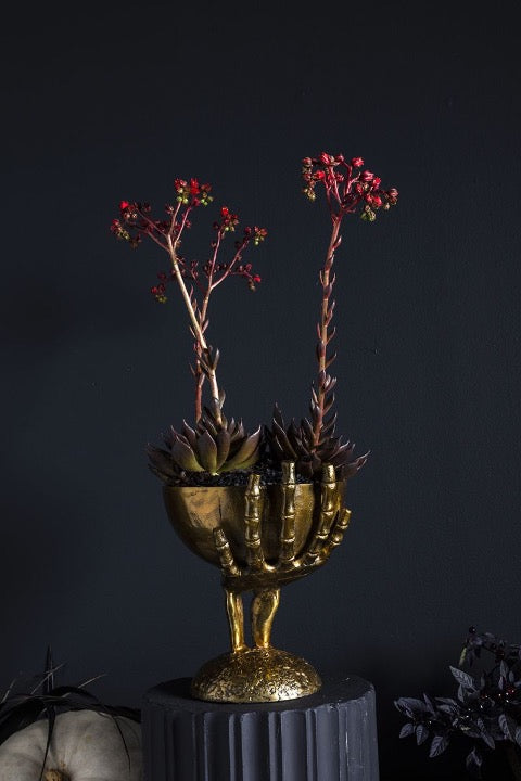 Metal skeleton hand holding bowl in brass finish with floral arrangement.