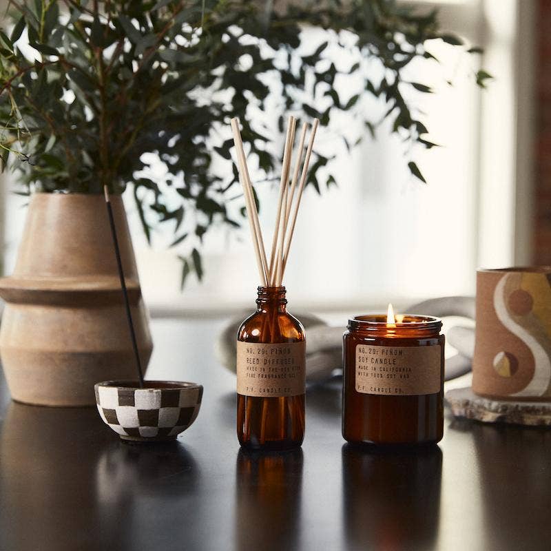 Lit Piñon candle beside a matching amber reed diffuser and a checkered incense bowl on a dark table