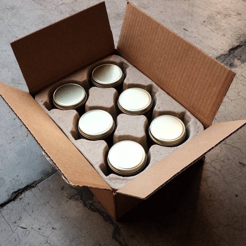 Six gold-lidded candle jars packed in a cardboard shipping box with a molded pulp insert