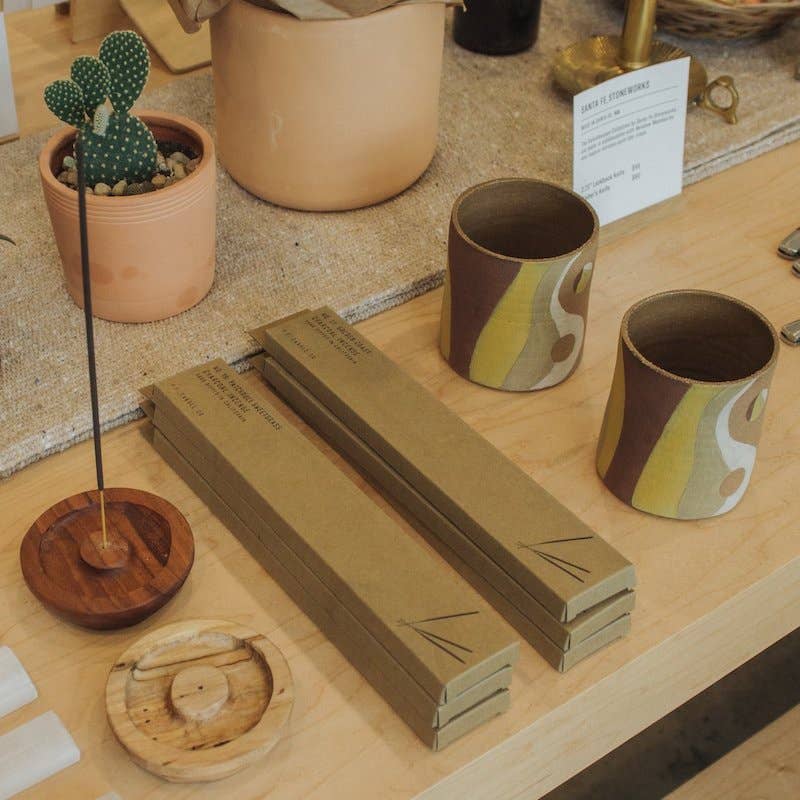 Incense boxes stacked on a wood shop table beside wooden incense holders, a cactus and striped cups