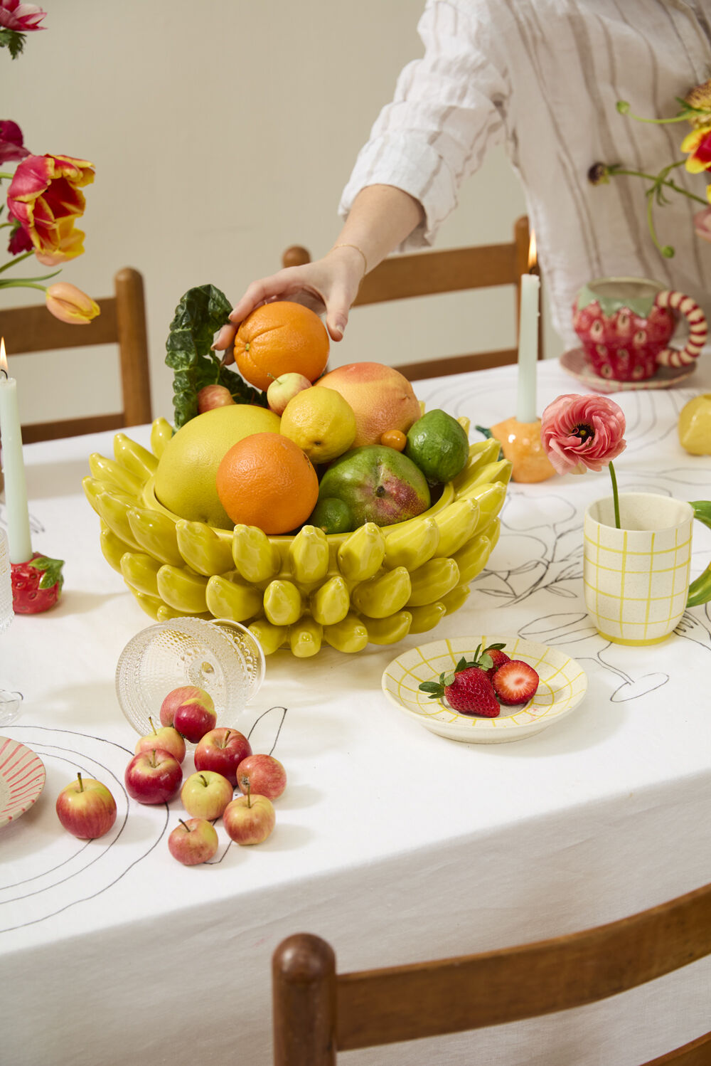 Banana bowl filled with oranges, apples and citrus on a white tablecloth as a hand adds an orange