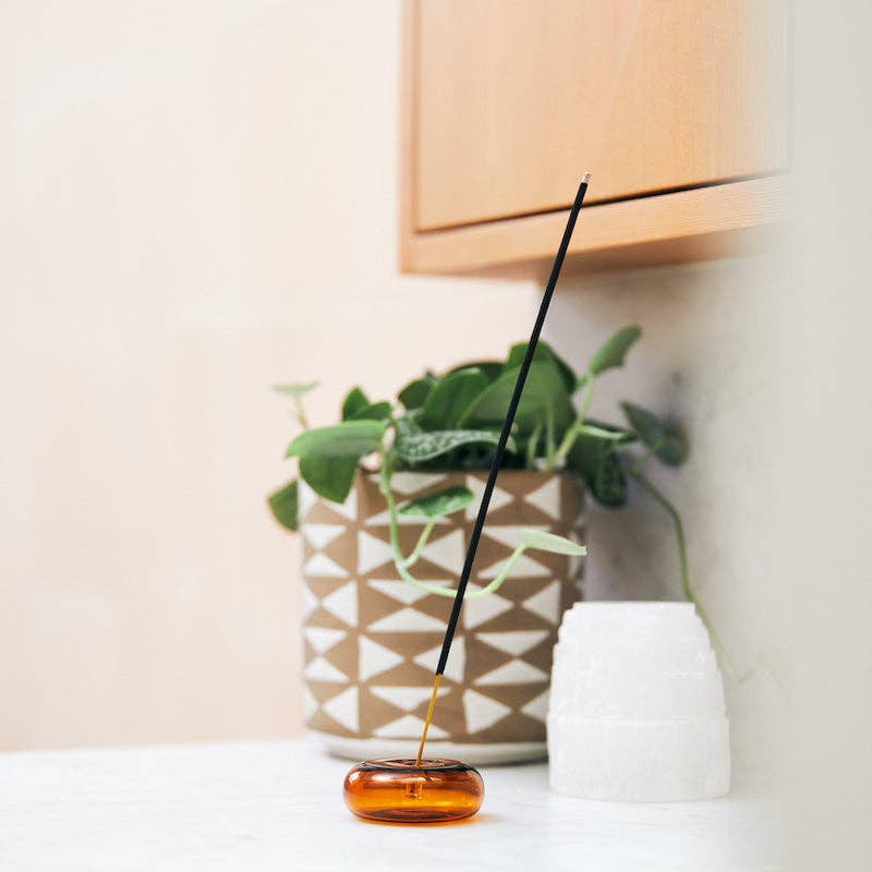 Amber & Moss incense stick standing in a round amber glass holder beside a potted plant on a white counter