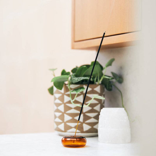 Amber & Moss incense stick standing in a round amber glass holder beside a potted plant on a white counter