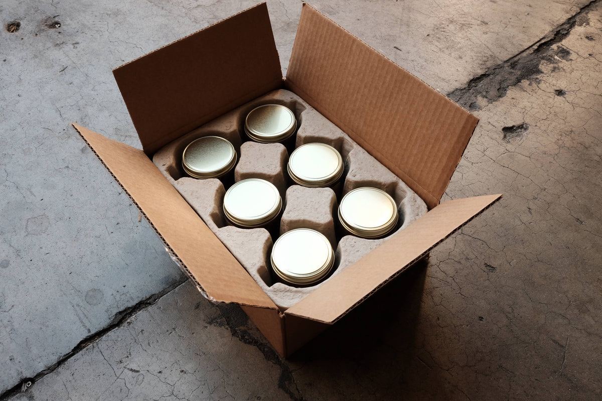 Open cardboard box with six gold-lidded candle jars nested in a molded pulp insert, seen from above