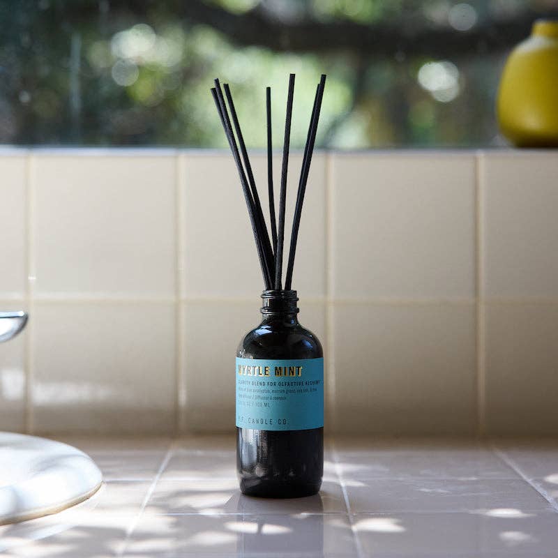 Myrtle Mint reed diffuser on a sunlit cream tile counter with greenery in the background