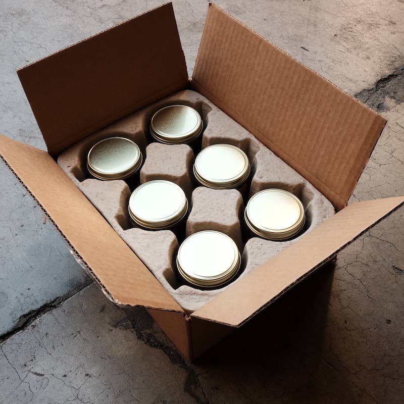 Open shipping box seen from above holding six candles with gold lids in a molded pulp insert