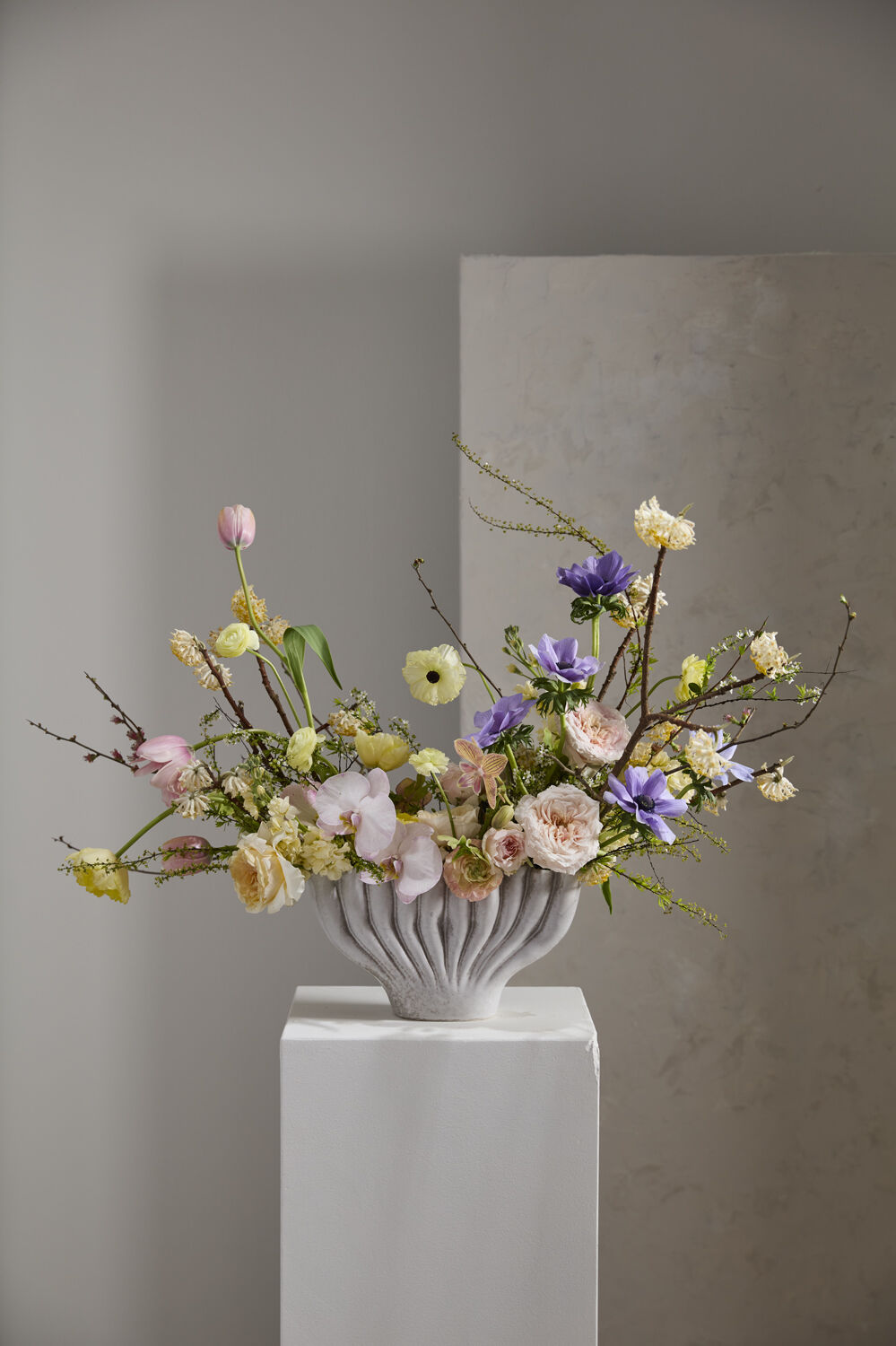 Clymene vase holding a loose arrangement of roses, orchids, tulips, and anemones on a white pedestal
