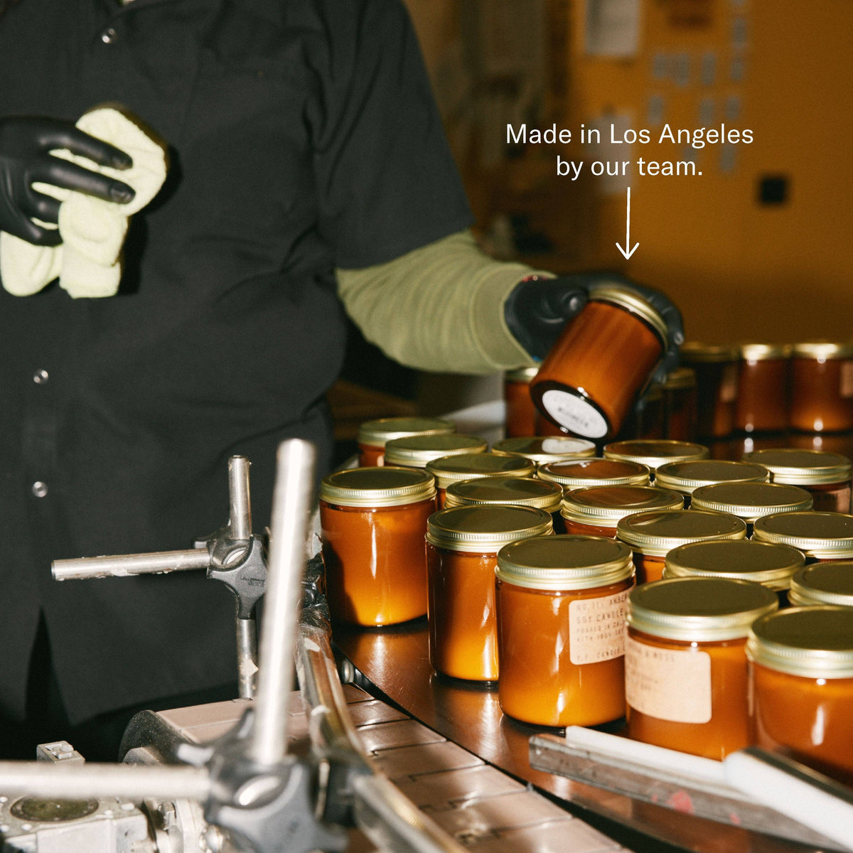 Gloved worker lifting a filled amber candle jar off a production line, captioned Made in Los Angeles by our team