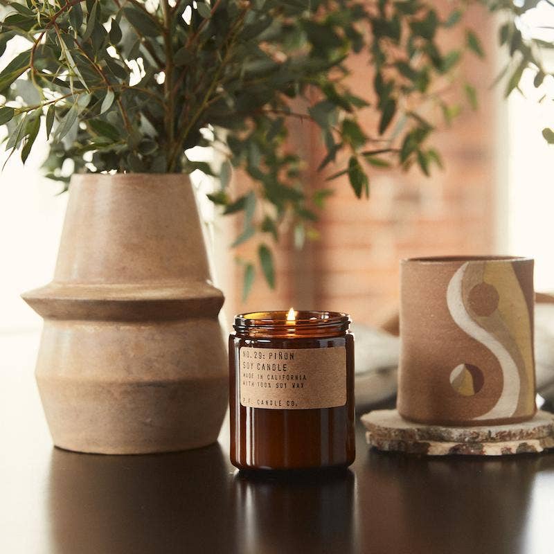 Lit Piñon candle in amber jar on a dark table between a tan vase of eucalyptus and a painted ceramic cup