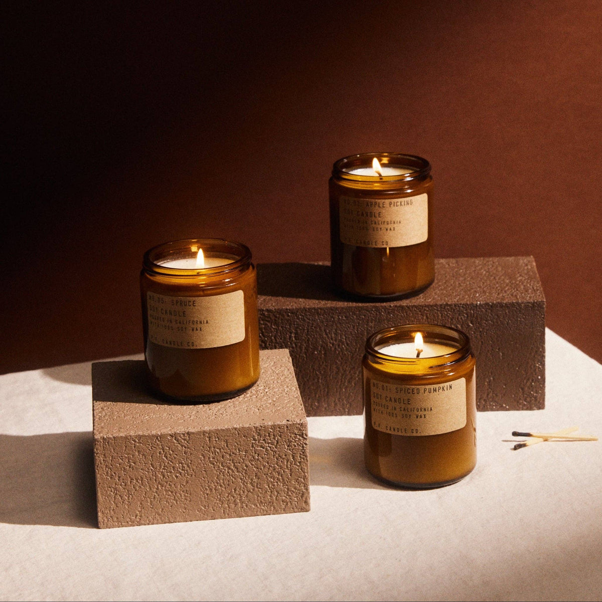 Lit Spruce candle with Apple Picking and Spiced Pumpkin candles on textured blocks