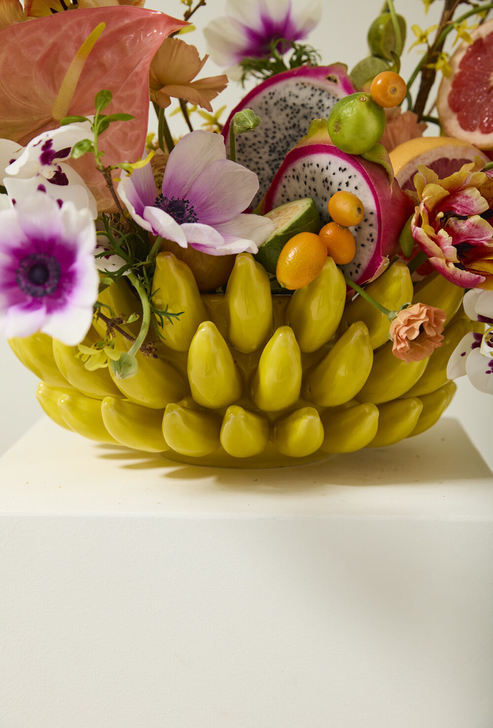 Close-up of yellow banana bowl overflowing with dragon fruit, kumquats, grapefruit and anemone flowers
