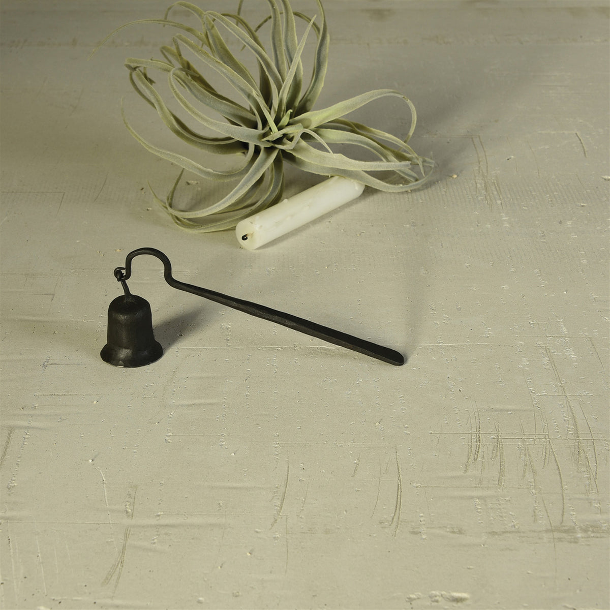 black hand-forged iron snuffer on concrete with candle and air plant in background