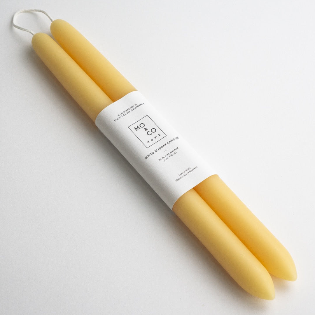 Two yellow beeswax tapers laid diagonally on white, showing tapered tips and the white paper label