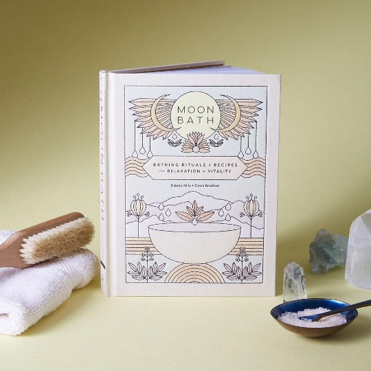 Moon Bath hardcover standing on a yellow surface with a bath brush, white towel, crystals and a bowl of salt