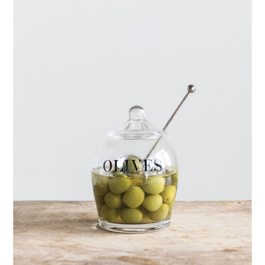 Olive jar filled with green olives in brine, steel spoon resting in the lid, on a rustic wood surface