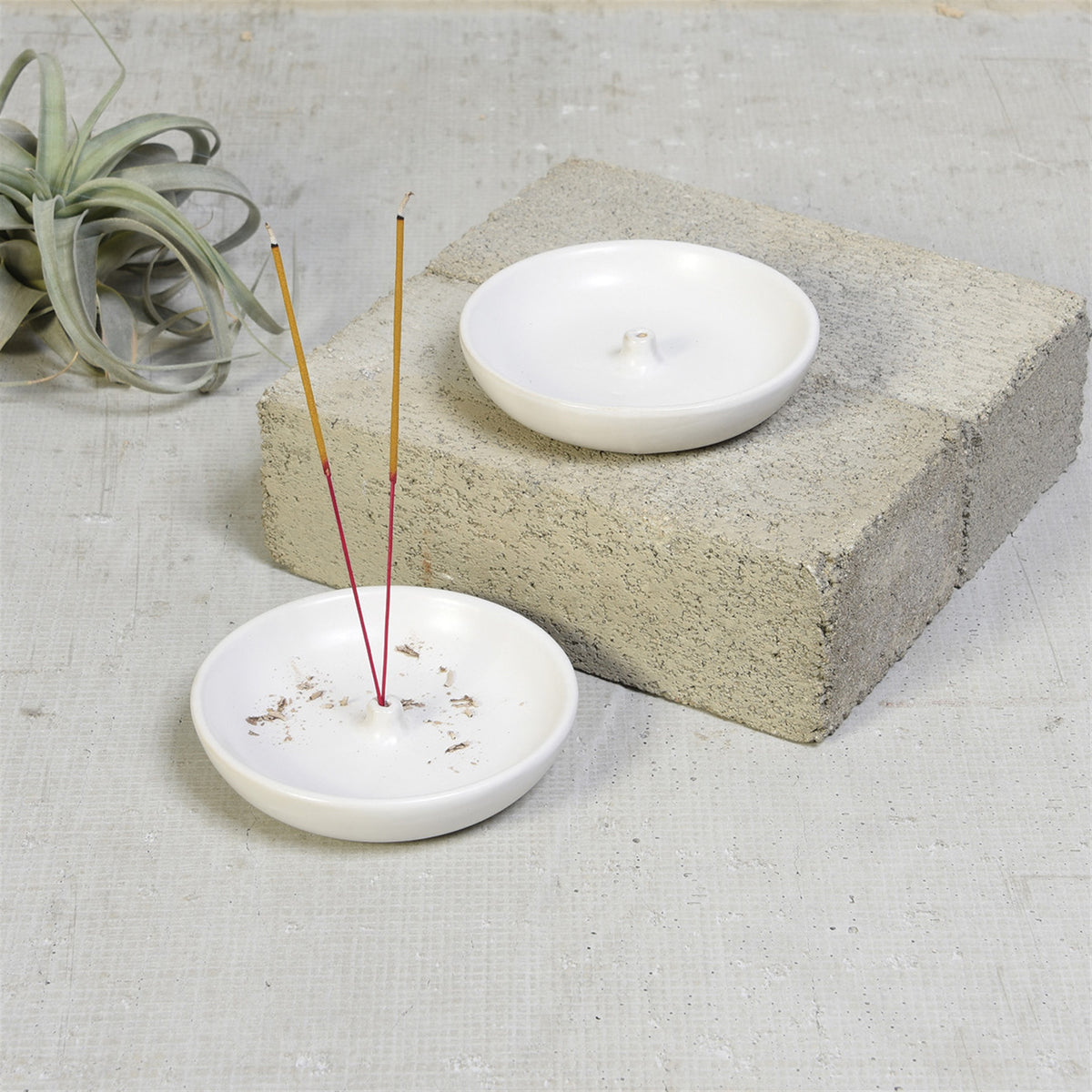 Two matte white Luna incense holders, one with two incense sticks, the other on a concrete block by an air plant