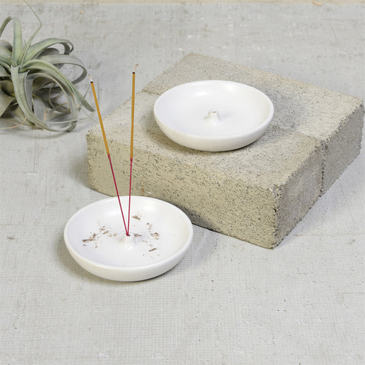 Two matte white Luna incense holders, one with two incense sticks, the other on a concrete block by an air plant