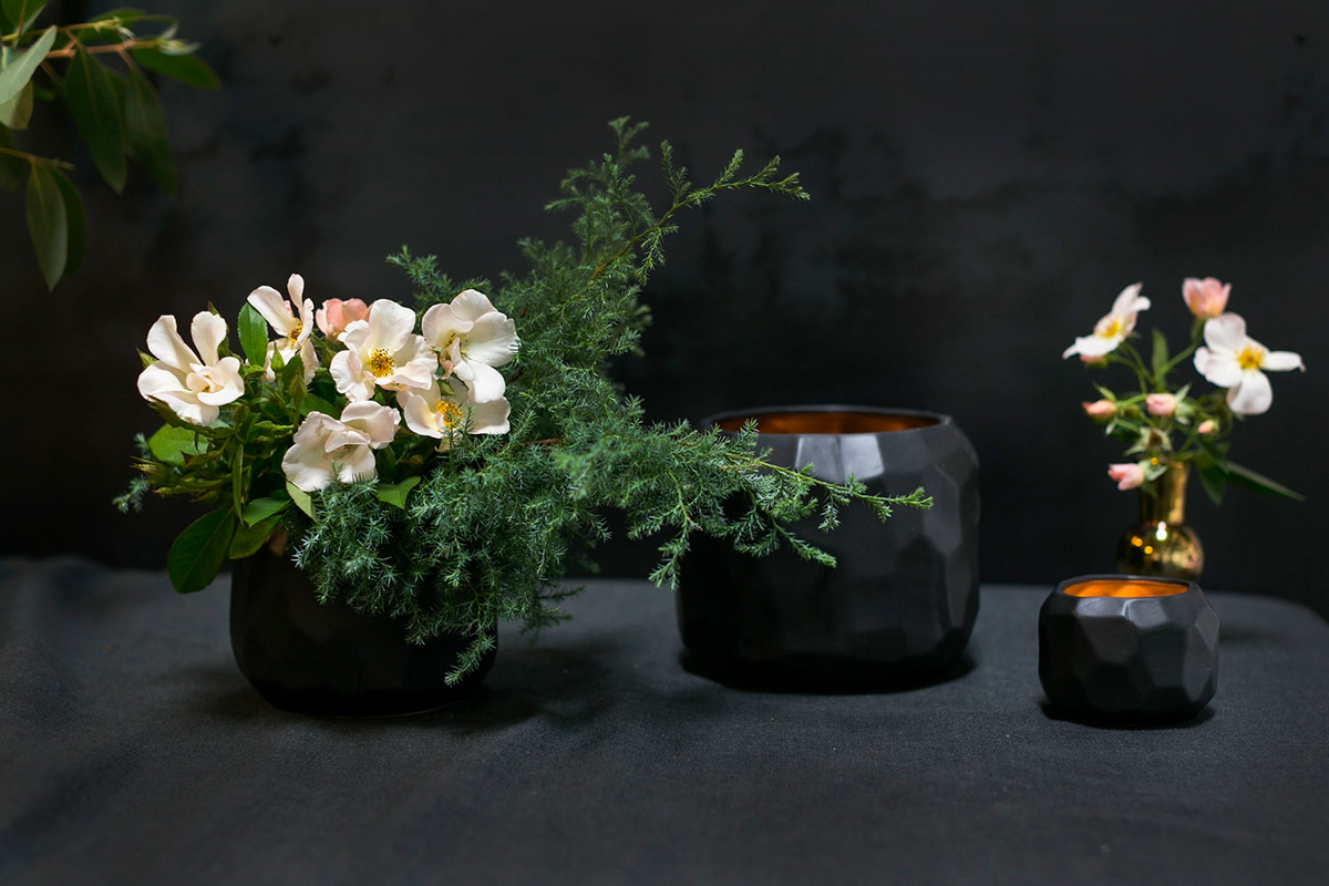 Three Miro pots on dark linen, one planted with white flowers and greenery, two with glowing amber interiors
