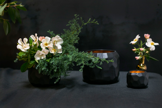 Three Miro pots on dark linen, one planted with white flowers and greenery, two with glowing amber interiors