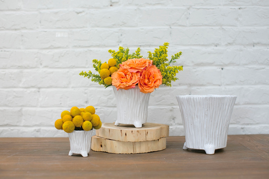 Mellie pots holding orange roses and yellow billy balls on a wooden table by a white brick wall