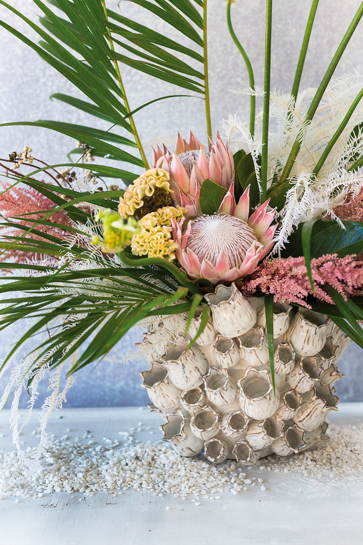 Marine vase holding a protea, palm fronds, and pink astilbe arrangement on white pebbles