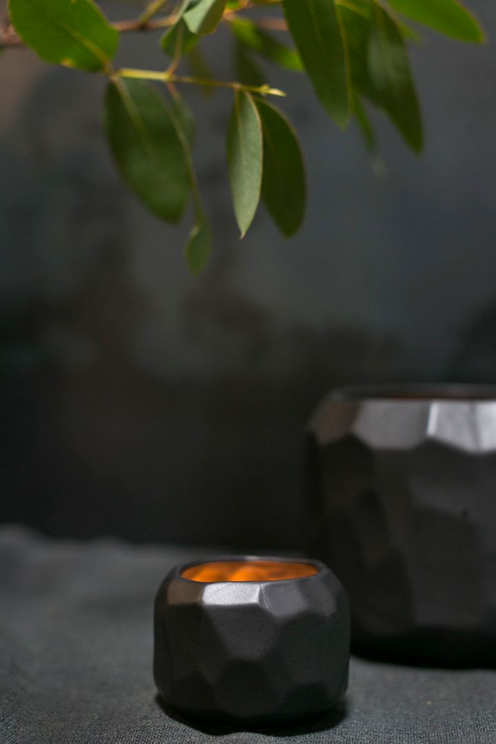 Small Miro pot with a glowing amber interior on dark linen beneath eucalyptus leaves, a larger pot behind